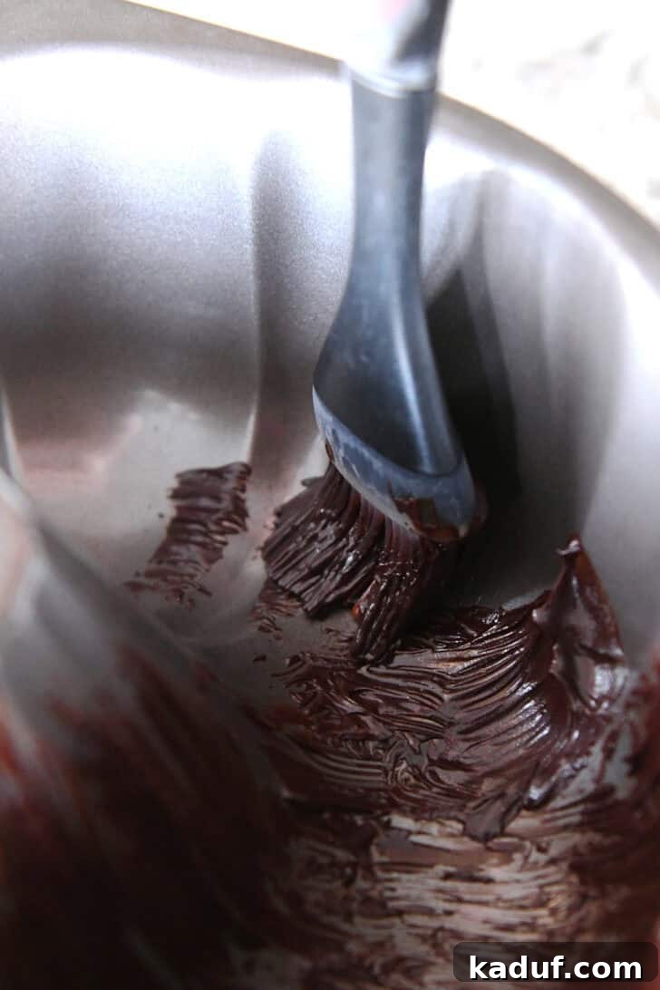 Velvet Chocolate Fudge Bundt 5 A pastry brush expertly applies a thin, even layer of chocolate butter mixture into the intricate fluted design of a gleaming metal bundt pan, ensuring a perfect cake release.