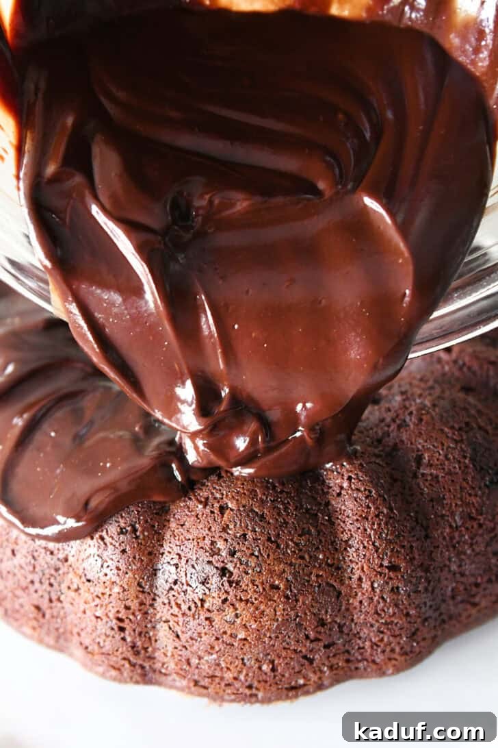 Velvet Chocolate Fudge Bundt 7 Thick, glossy chocolate ganache being poured from a clear glass bowl onto a baked chocolate bundt cake, creating luscious, elegant drips down the fluted sides.