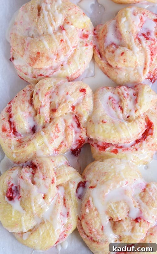 Overnight Strawberry Cheesecake Rolls 2 Six, baked strawberry cream cheese rolls on a piece of parchment paper drizzled with a white glaze.
