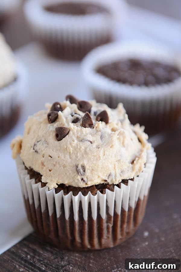 Decadent Fudge Brownie Cupcakes 4 A close-up of a chocolate fudge brownie cupcake, generously topped with a towering scoop of rich, creamy cookie dough frosting, set against a blurred background.