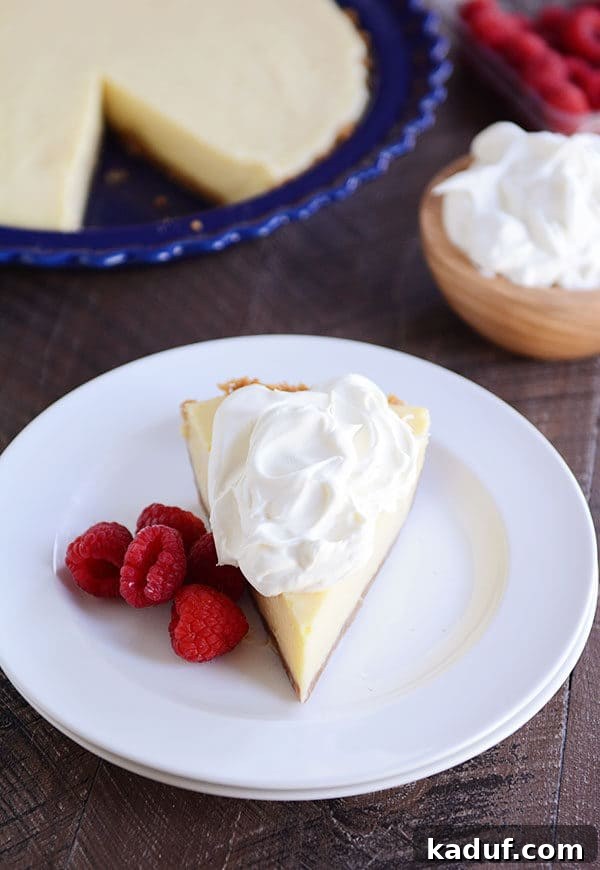 A beautifully presented slice of creamy lemon pie, adorned with a swirl of light whipped cream and several fresh, vibrant raspberries on a pristine white plate. In the blurred background, the remainder of the pie sits invitingly in a rustic blue pie dish.
