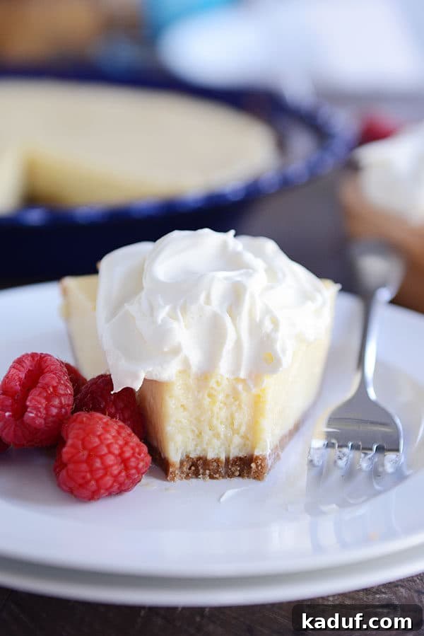 A close-up view of a single slice of lemon pie on a white plate, with a delectable bite already taken. The smooth, pale yellow filling and distinct graham cracker crust are visible, generously topped with whipped cream and garnished with fresh raspberries.