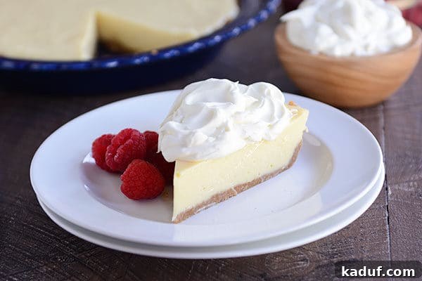 An overhead shot showcasing a luscious slice of lemon pie, crowned with fluffy whipped cream and scattered fresh raspberries, elegantly presented on a white dessert plate. The rest of the golden-crusted pie waits in a blue ceramic pie dish in the soft background.