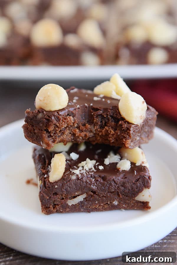 Hawaiian Chocolate Macadamia Brownies 4 A stack of two chocolate macadamia nut brownies, one with a tempting bite taken out, revealing the soft, fudgy interior and crunchy nut pieces.