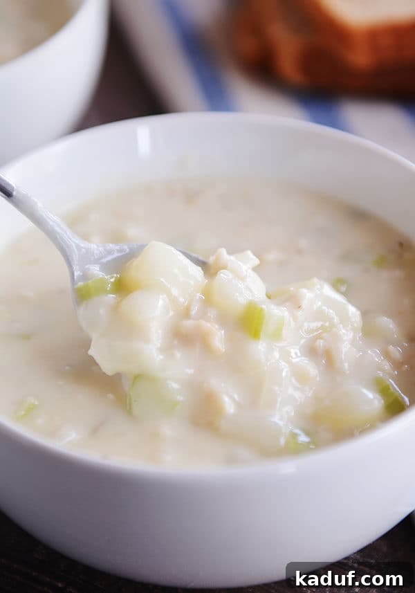 Our Signature New England Clam Chowder 2 A spoon taking a big bite of creamy New England clam chowder out of a white bowl, showing tender clams and potatoes.