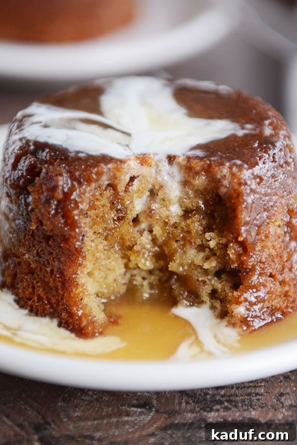 Gloriously Sticky Toffee Pudding Cake 6 A small brown cake, with a bite taken out, covered with sticky toffee sauce and cream on a white plate. Shows the moist interior of the cake.