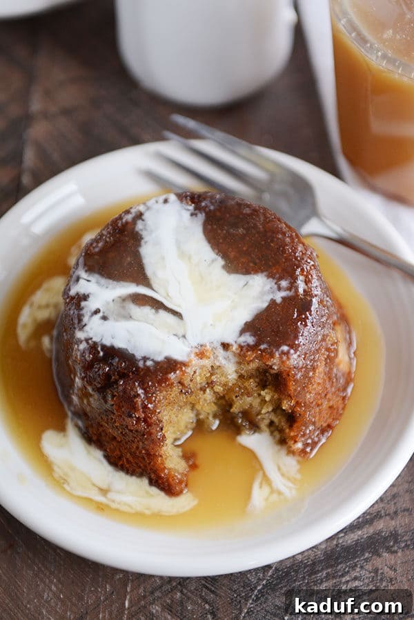 Gloriously Sticky Toffee Pudding Cake 7 A small brown cake, with a bite taken out, covered with sticky toffee sauce and cream on a white plate. Close up of the cake's texture and moistness.