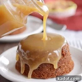 A small, brown cake on a white plate, with light brown sticky toffee sauce being poured over the top.