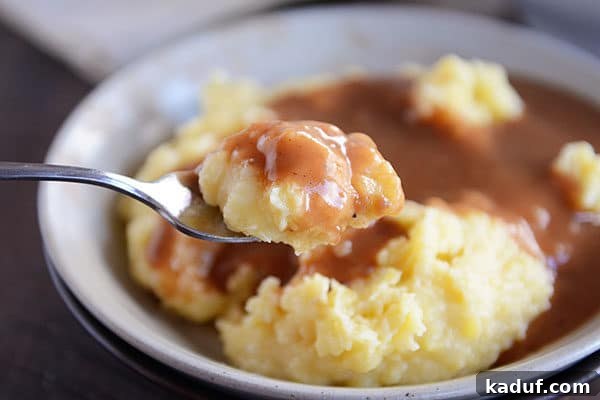 Velvety Instant Pot Mashed Potatoes 8 A spoonful of mashed potatoes and brown gravy, with a bowl full of mashed potatoes and brown gravy in the background.