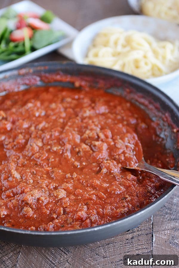 Whip Up a Quick Homemade Spaghetti Sauce Tonight 3 A vibrant red, meaty spaghetti sauce simmers in a skillet, with cooked spaghetti noodles and a fresh green salad blurred in the background, hinting at a complete meal.