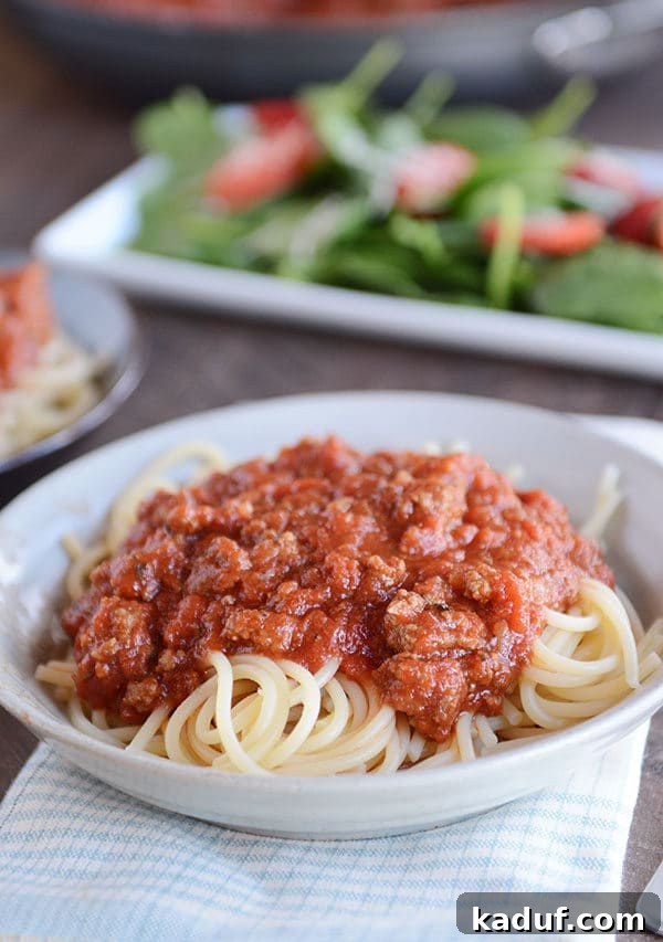 Whip Up a Quick Homemade Spaghetti Sauce Tonight 4 A white bowl brimming with cooked spaghetti noodles, generously covered in rich, meaty red spaghetti sauce, with a fresh green salad serving as a wholesome backdrop.