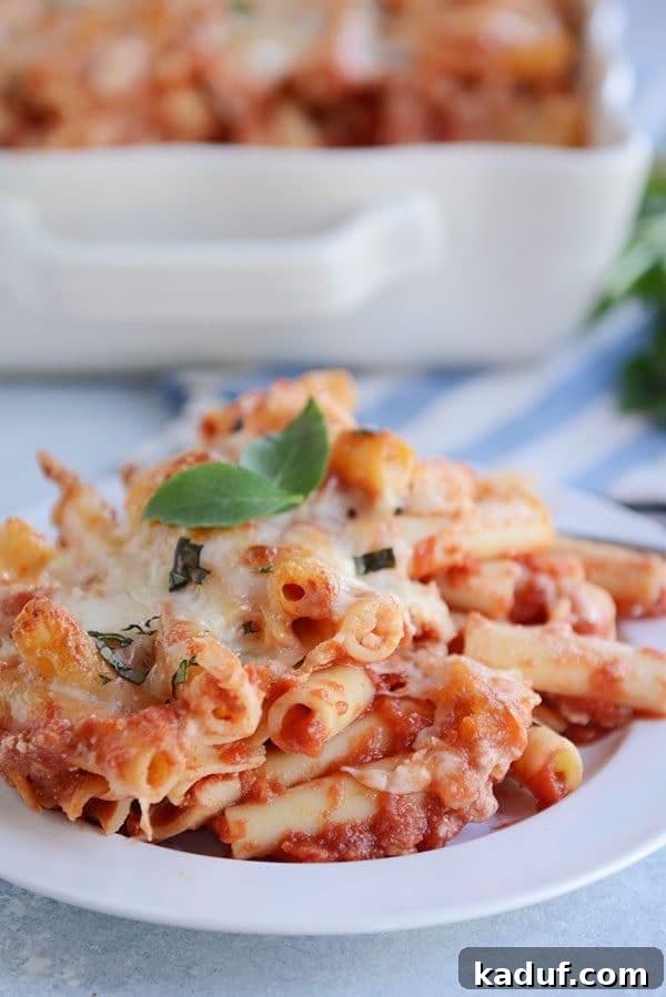 Hearty Make-Ahead Cheesy Baked Ziti 6 A white plate of cheesy baked ziti with red sauce and two basil leaves on top.
