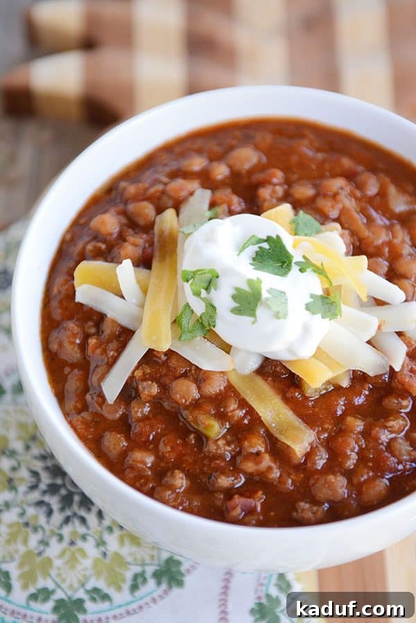 A white bowl full of sour cream and cheddar cheese-topped lentil beef chili, garnished with fresh herbs.