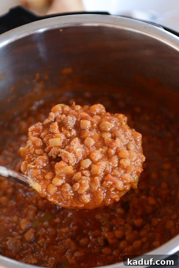 A metal ladle scooping rich, thick lentil and beef chili from a large pot, steam rising.
