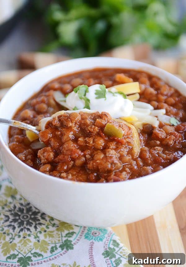 A spoon digging into a white bowl of hearty lentil and beef chili, showcasing its rich texture.