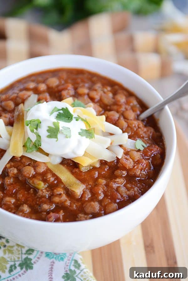 A vibrant white bowl of lentil chili, beautifully garnished with a dollop of sour cream, shredded cheddar cheese, and fresh parsley.