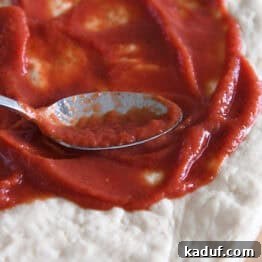 A metal spoon spreading vibrant pizza sauce on flattened, uncooked pizza dough.