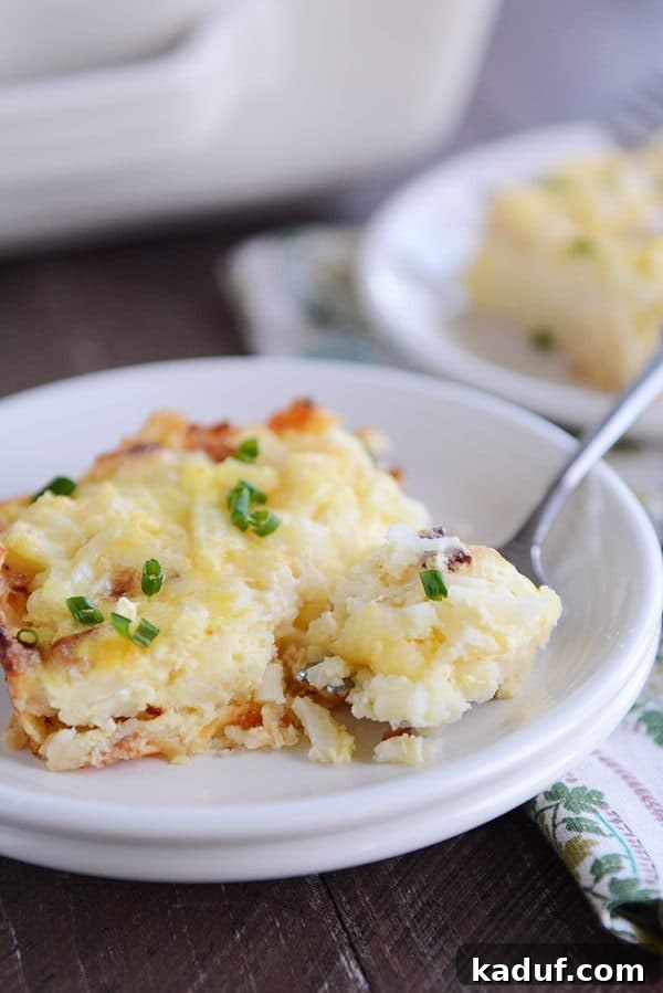 Rich Cheesy Hash Brown and Egg Overnight Bake 3 A piece of cheesy egg and hash brown casserole on a white plate, with a fork taking a bite out.