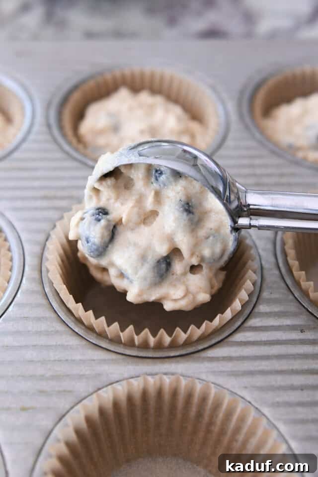 A hand expertly scooping wholesome muffin batter into a prepared muffin tin using a cookie scoop, ensuring even portions.