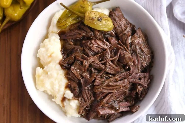 White shallow bowl with mashed potatoes, two pepperoncini, and shredded beef.