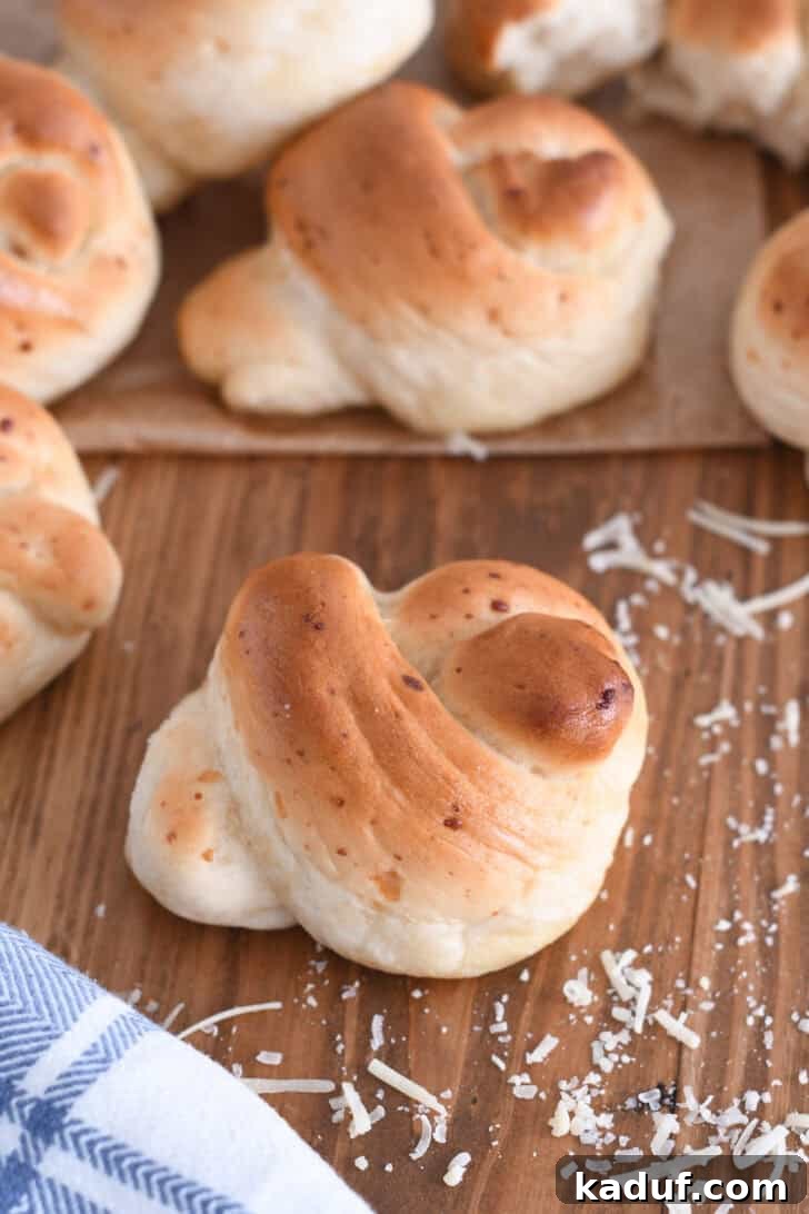 Quick Cheesy Parmesan Knots 2 A perfectly baked Parmesan breadstick knot, garnished with shredded Parmesan cheese, resting on a rustic wooden board beside a white and blue patterned cloth, highlighting its golden crust and fluffy texture.