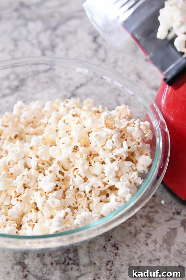 Gooey Peanut Butter Popcorn Bliss 5 An air popper vigorously popping popcorn kernels into a clear glass bowl, filling it with light, fluffy white popcorn.