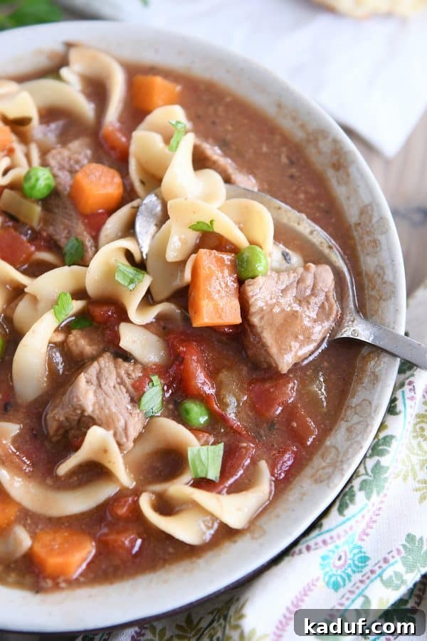 Hearty Slow Cooker Beef and Veggie Noodle Soup 2 Top down view of slow cooker beef vegetable noodle soup with spoon in gray ceramic bowl, garnished with fresh herbs.