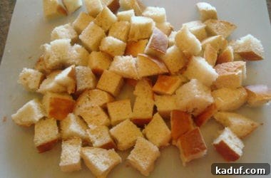 A large pile of freshly cut one-inch bread cubes sits on a white cutting board, awaiting seasoning and baking to transform into crispy croutons.