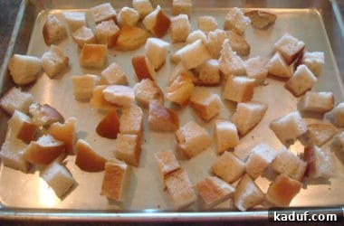 Unbaked bread cubes are evenly spread in a single layer on a metal cookie sheet, ready for seasoning and toasting in the oven.