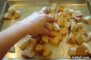 Bread cubes on a cookie sheet are generously drizzled with olive oil and tossed by hand to ensure even coating, essential for crispy, flavorful croutons.