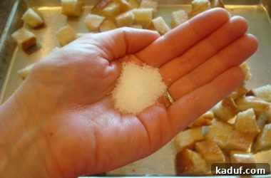 A hand sprinkles coarse salt evenly over a pan of olive oil-coated bread cubes, enhancing flavor and promoting crispness.