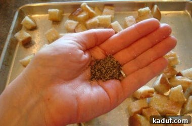 Freshly cracked black pepper is generously sprinkled by hand over a sheet pan of seasoned bread cubes, adding a pungent aroma and bold flavor.