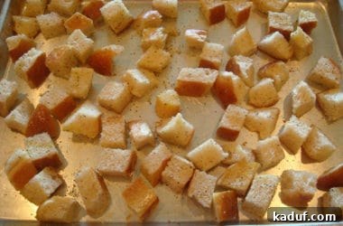 Seasoned bread cubes are arranged on a metal cookie sheet, ready to be baked in a hot oven until golden brown and crispy.