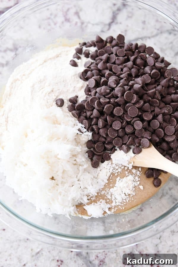 Ultimate Oatmeal Chocolate Chip Coconut Cookie Bars 7 Flour, coconut and chocolate chips in glass bowl with batter ingredients.