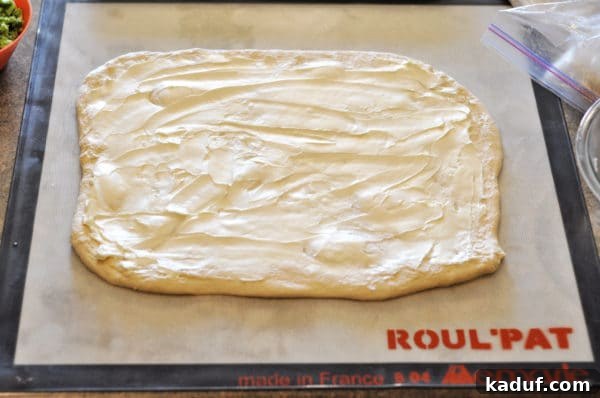 Spread The rolled-out rectangular bread dough is generously and evenly brushed with a creamy, pale yellow mixture of softened butter and cream cheese, covering the entire surface.