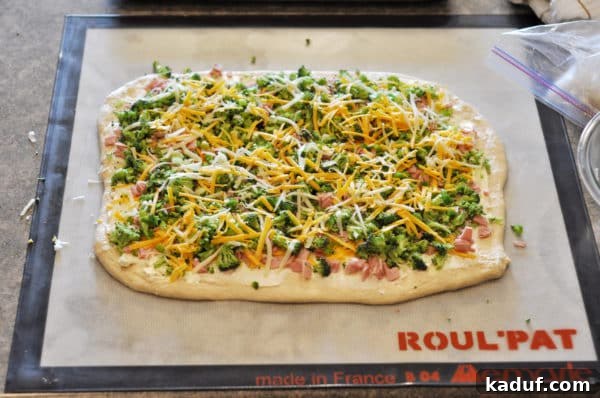 All Toppings Uncooked bread dough, covered with a vibrant and generous layer of finely chopped pink ham, small green florets of broccoli, and a golden scattering of shredded cheese, ready to be rolled.