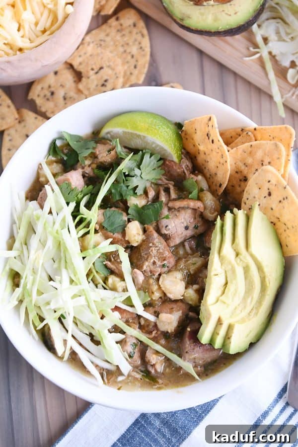 Easy Slow Cooker Posole 2 Top down view of slow cooker posole in white bowl with shredded cabbage, sliced avocado, tortilla chips and lime wedge.