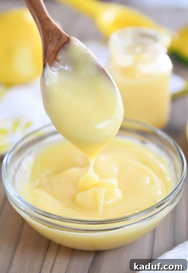 Easy Instant Pot Lemon Curd 2 Wooden spoon dropping freshly made lemon curd into a clear glass bowl, showcasing its smooth texture and vibrant yellow color.