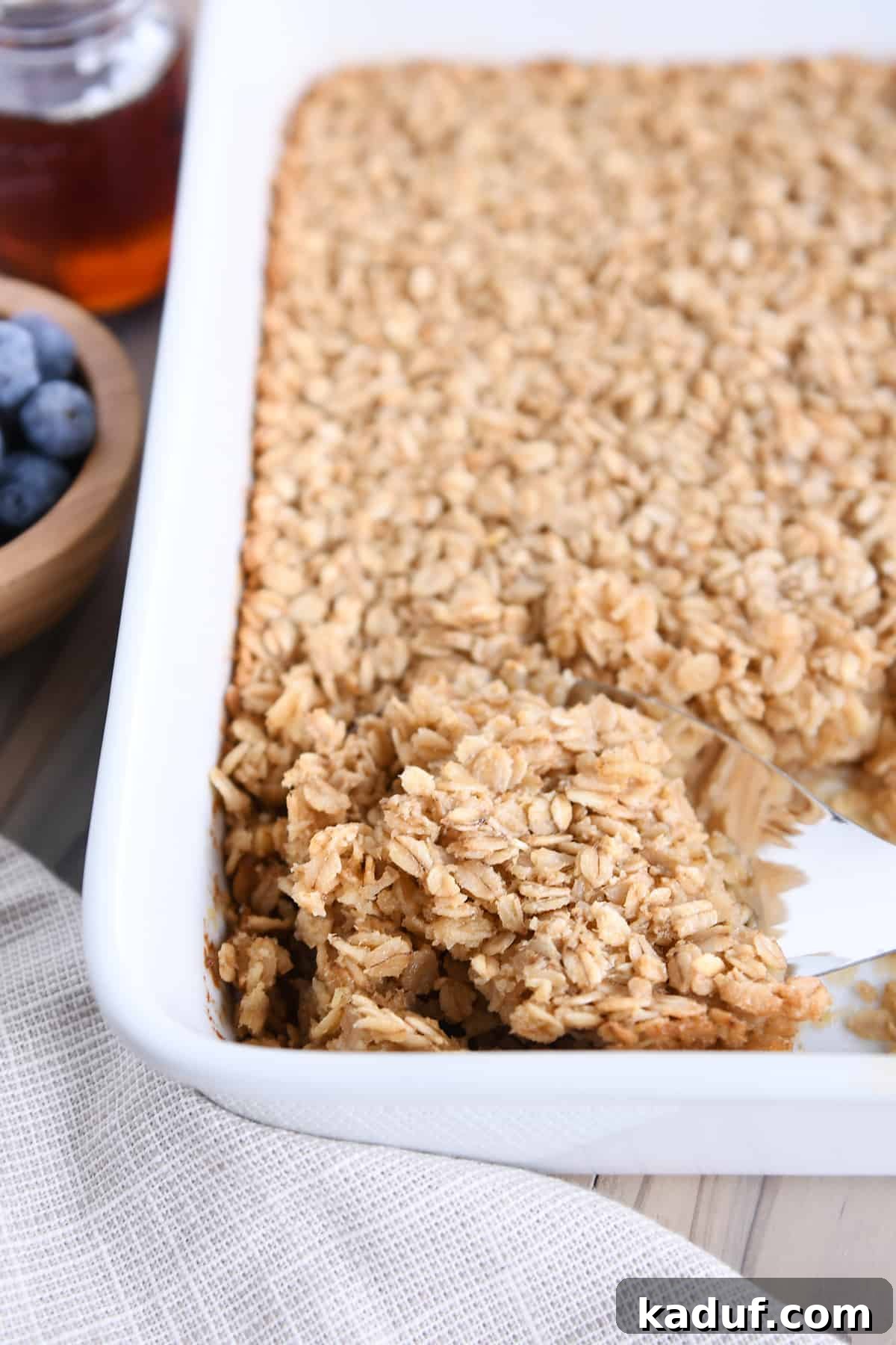 Effortless Amish Overnight Baked Oats 6 A serving spoon scoops a generous portion of freshly baked Amish oatmeal from a white baking dish, showcasing its perfect golden-brown edges and inviting texture.