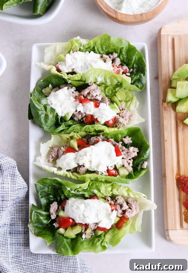 Zesty Greek Lettuce Boats 2 Four beautifully arranged Greek lettuce wraps on a white tray, accompanied by fresh cucumbers, ripe tomatoes, and a small bowl of creamy feta mint dressing.