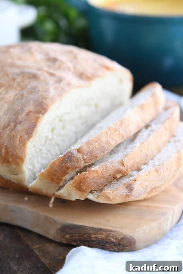 Pratik Yoğurmasız Köy Ekmeği 2 A freshly baked loaf of easy no-knead peasant bread resting on a rustic wood cutting board, with three inviting slices already cut and ready to enjoy.