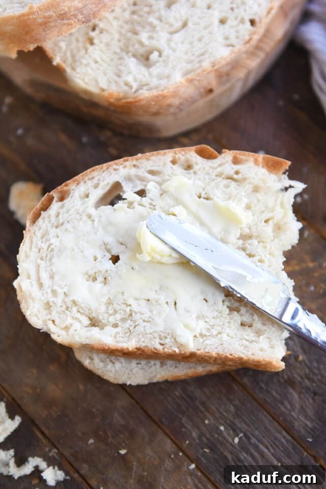 Pratik Yoğurmasız Köy Ekmeği 3 A hand gently spreading butter onto a half slice of the freshly baked no-knead peasant bread with a butter knife, highlighting its soft texture.