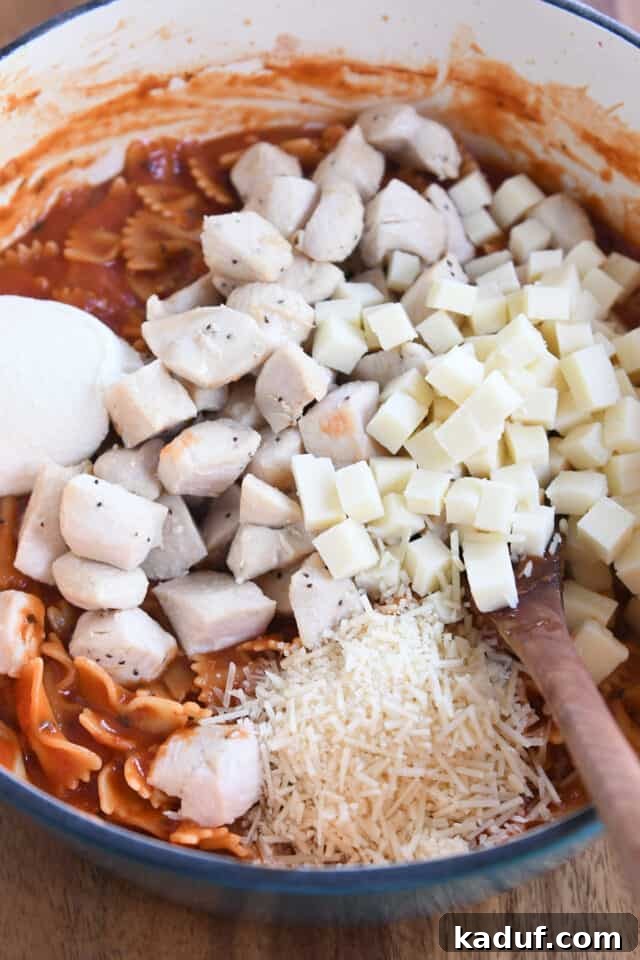 Effortless One-Pot Chicken Lasagna 5 Mozzarella cubes, cooked chicken, parmesan cheese, and ricotta cheese mixed with bowtie pasta in a cast iron pot, showcasing the final step before serving.