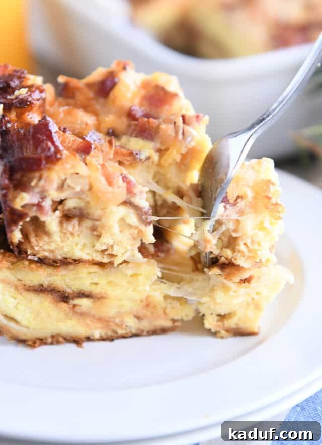 Overnight Cheesy Bacon Croissant Strata 7 Close-up of a fork taking a bite from the breakfast casserole on a white plate, showing melted cheese stretching.