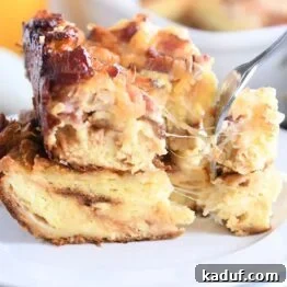 Overnight Cheesy Bacon Croissant Strata 8 cheese strings pulling as fork takes bite from breakfast casserole on white plate
