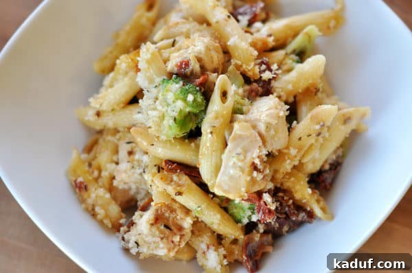 Smoky Mozzarella Chicken and Broccoli Penne Bake 10 Top-down view of a white rectangular dish brimming with baked penne, featuring tender chicken, green broccoli florets, and red sun-dried tomatoes, all nestled in a creamy sauce and topped with golden crumbs.