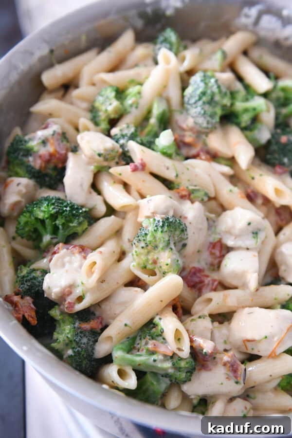 Smoky Mozzarella Chicken and Broccoli Penne Bake 5 A large stainless steel pot filled with a rich mixture of penne pasta, vibrant green broccoli florets, sun-dried tomatoes, and creamy white sauce, before being transferred to a baking dish.