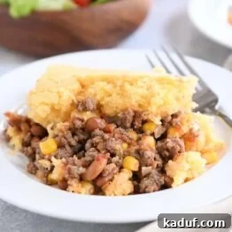 A perfect portion of cornbread topped beef and bean casserole on a white plate with a fork.
