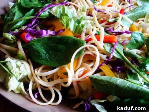 Ginger Sesame Noodle Salad 2 A large white bowl filled with a colorful Asian noodle salad, featuring linguine noodles, red and yellow bell peppers, and fresh spinach.