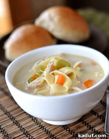 Heartwarming Chicken Noodle Soup 2 A white bowl full of classic homemade chicken noodle soup with golden rolls in the background, set on a rustic wooden surface.